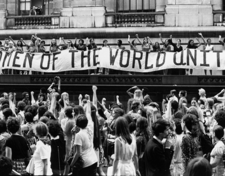 Crowd of people protesting below a large banner that reads 'Women of the World Unite!'