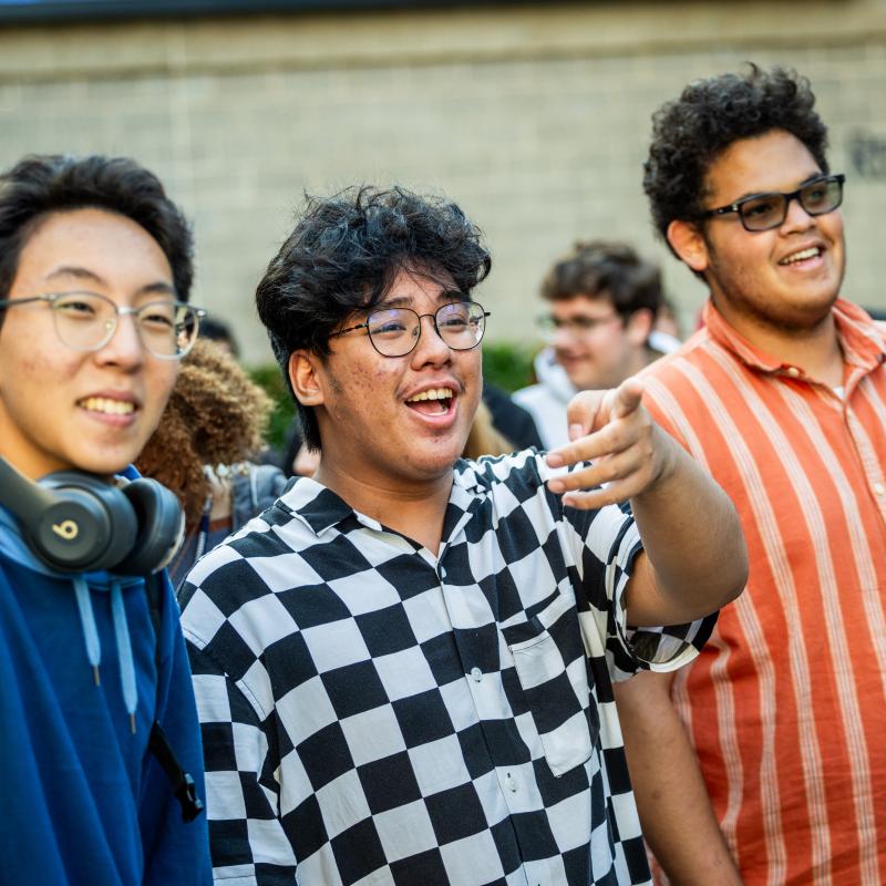 Asian students smile and point toward an off-camera activity while gathered together at an outdoor campus event.
