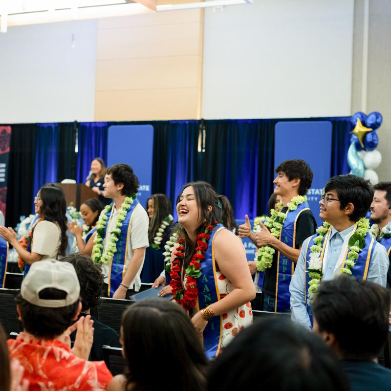 Students wearing ceremonial stoles and flower leis celebrate their achievements as family, friends, and classmates applaud during a campus recognition ceremony.