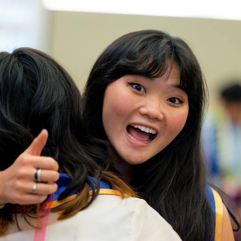 A student gives a thumbs-up while hugging a friend at a campus event and smiling enthusiastically toward the camera.