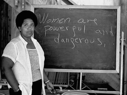 A black women in front of a chalk board that has the words "Women are powerful and dangerous"