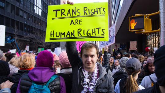 Person holding a bright yellow sign that reads “Trans Rights Are Human Rights” at a crowded protest in a city street.